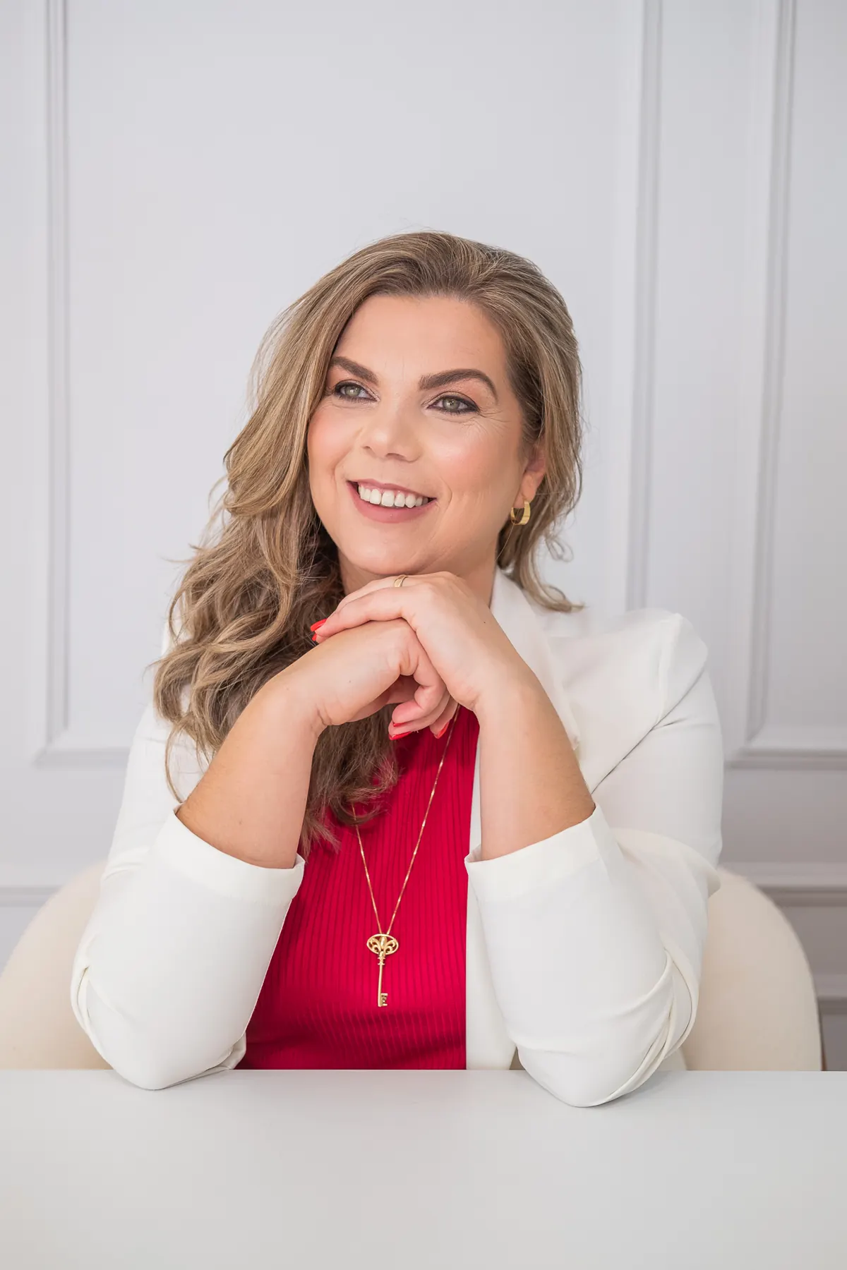 Lucinda, astróloga, de blazer branco e colar de chave, com as mãos apoiadas no queixo