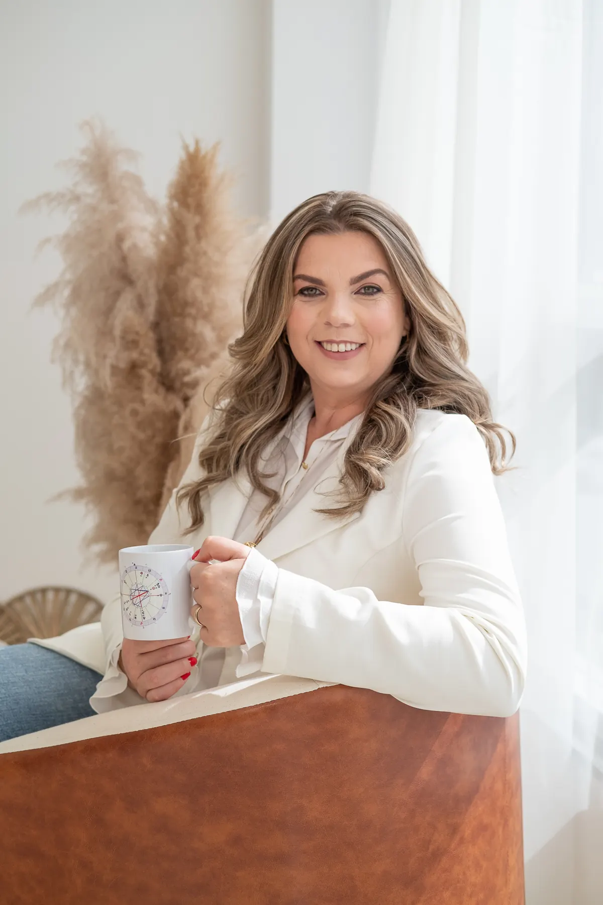 Lucinda sentada em poltrona, segurando uma caneca, em ambiente claro com plumas secas