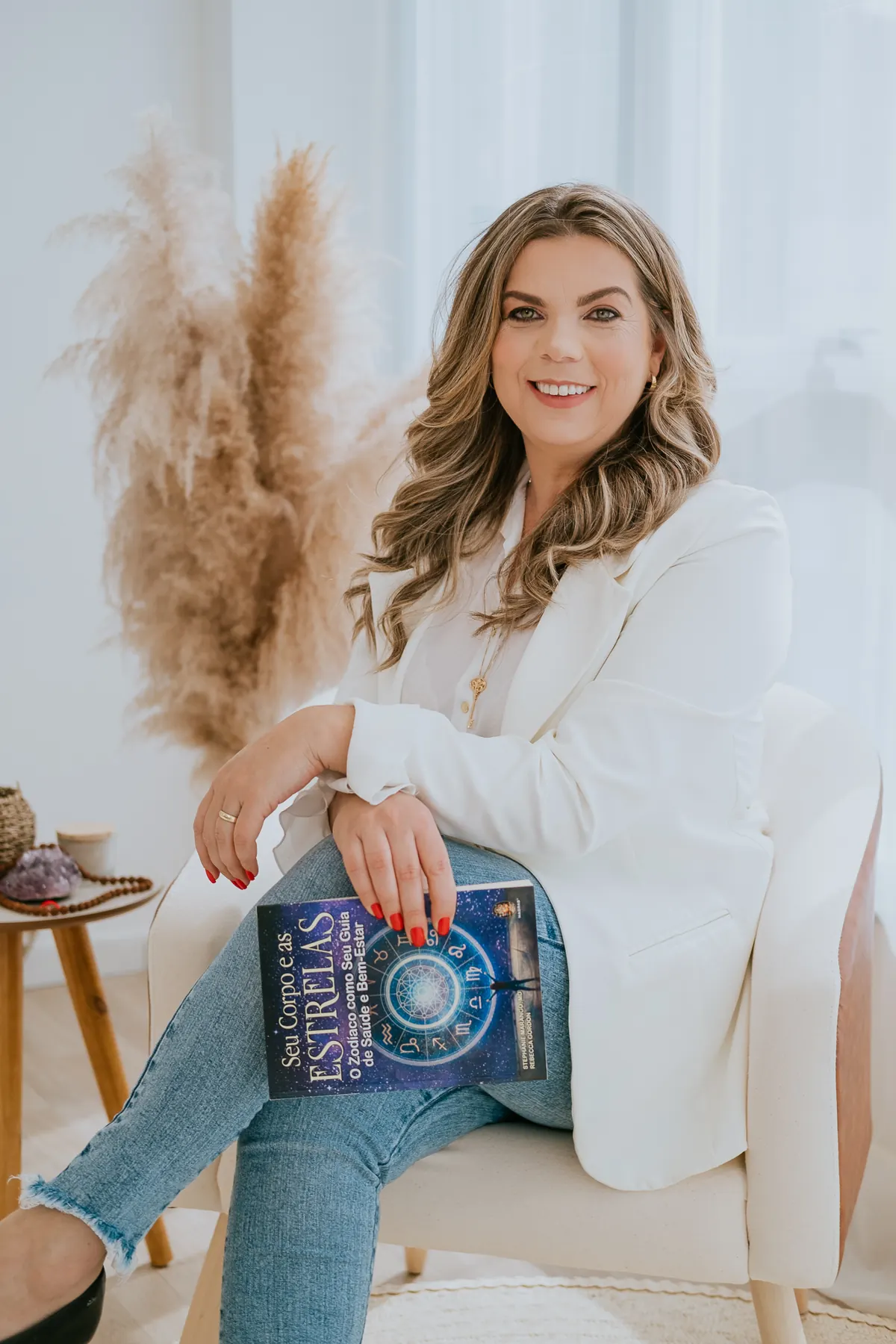 Lucinda sentada com um livro de astrologia no colo, em ambiente claro com plumas secas